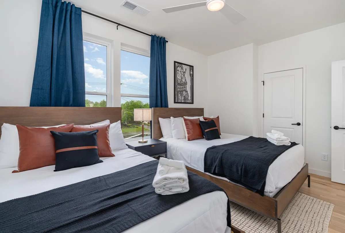 A spacious, light-filled bedroom with two beds dressed in decorative pillows, large windows with flowing curtains, a nightstand with a lamp, and tasteful artwork on the wall. The room exudes a relaxed and inviting atmosphere, ideal for both rest and socializing.