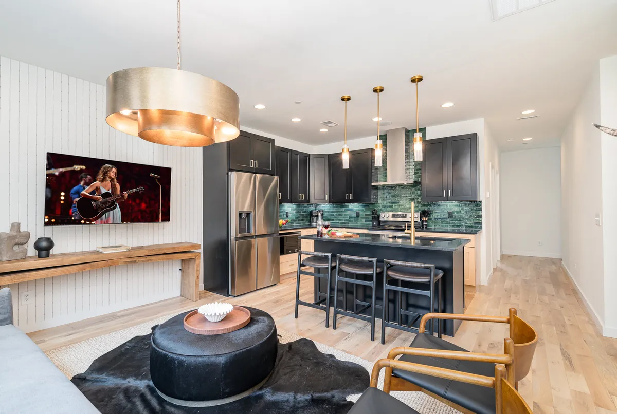 A contemporary kitchen and lounge space with a kitchen island, pendant lights, a TV displaying a musician in performance, and modern decor. The wooden bench and black accent pieces add sophistication, while the overall atmosphere is lively and set up for entertaining.