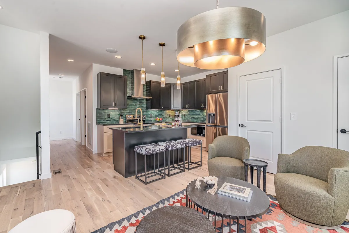 A cozy, contemporary kitchen with a green tile backsplash, pendant lighting, kitchen island with seating, and adjacent lounge area. The space invites both cooking and casual gatherings.