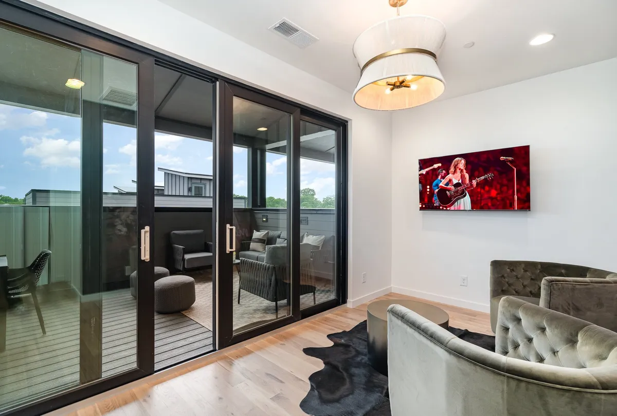 Large sliding glass doors dominate the scene, framing a stylish interior with a modern chandelier and flat-screen TV, suggesting a bright, open setting with easy access to outdoor spaces. The natural light and the potential for an outdoor view emphasize the connection to fresh air and open skies.