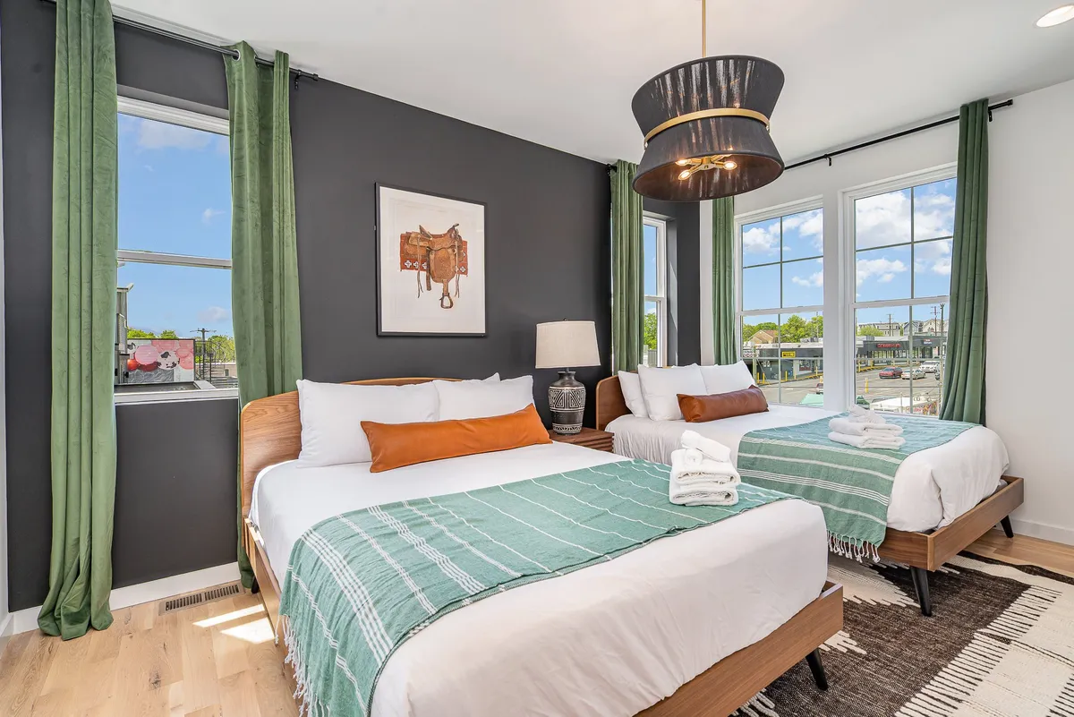 A comfortable bedroom featuring two beds with decorative throws, large windows adorned with green curtains, modern artwork, and a stylish pendant light. The room balances comfort and style, promoting both relaxation and social interaction.