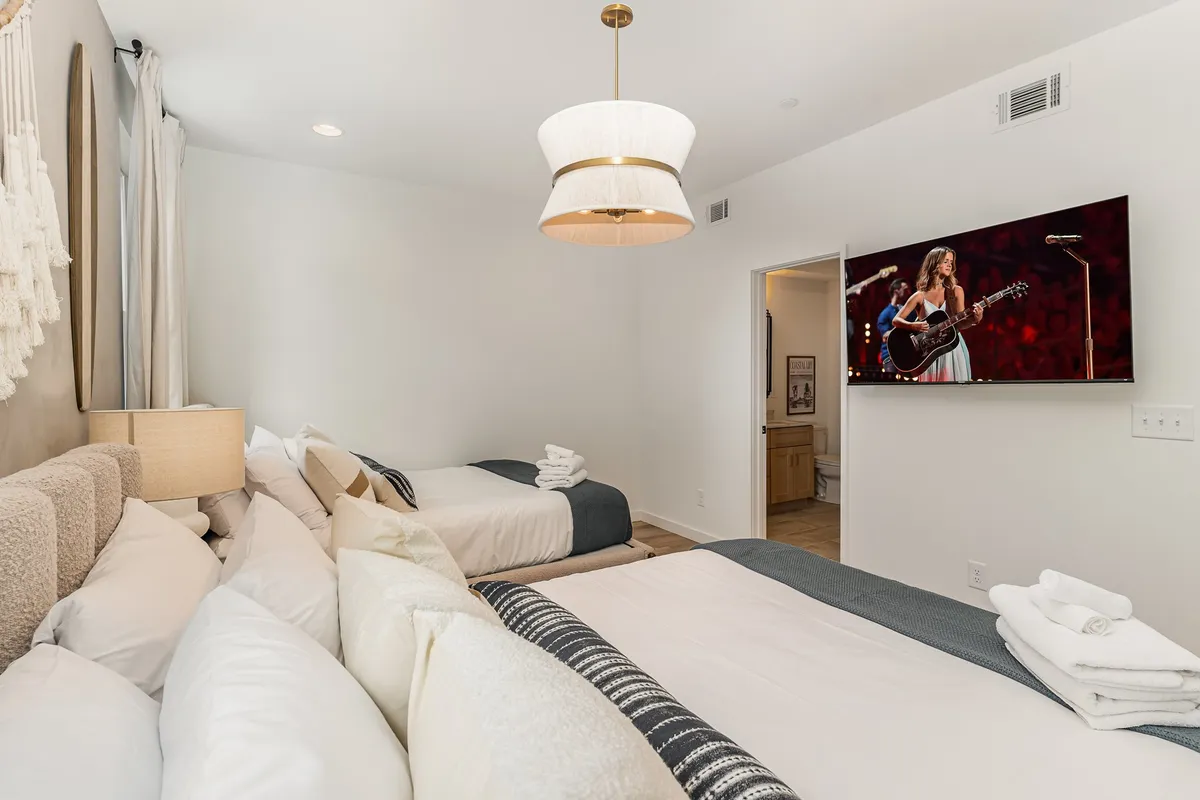 A comfortable bedroom with two beds and a TV screen featuring a performance, complemented by a pendant light fixture. The layout is welcoming and perfect for shared experiences, whether relaxing after an event, watching a concert, or socializing.
