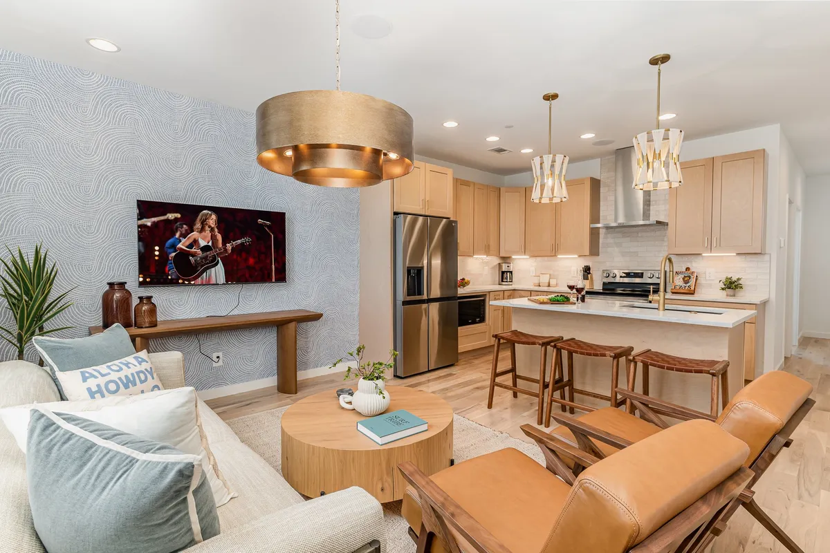 A cozy, open-concept living area featuring a plush sofa with decorative pillows, a wooden coffee table, a kitchen island with bar stools, and a statement wall art of a musician. Hanging light fixtures add warmth and a sense of gathering, while the spacious layout encourages socializing and entertainment.