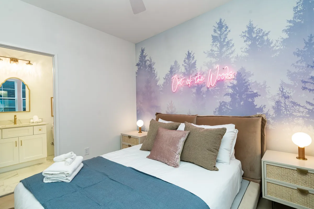 A tranquil bedroom with textured bedding, wooden nightstands, and a glowing neon sign reading 'Out of the Woods.' The scene is cozy and serene, suggesting a nature-inspired escape where guests can unwind after outdoor activities.