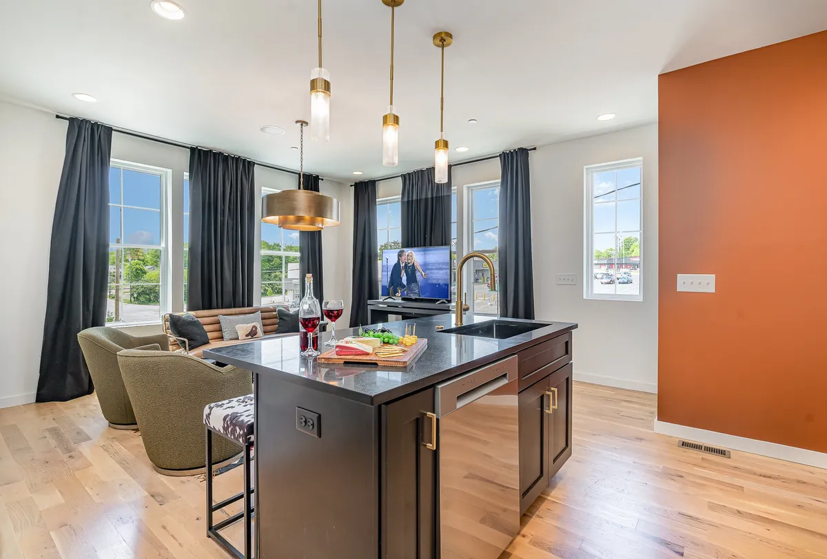A bright, open-plan living and kitchen space featuring a kitchen island, comfy sofa seating, a television, and large windows letting in abundant natural light. The space is arranged for both social gatherings and relaxed family moments, suitable for guests of all ages.